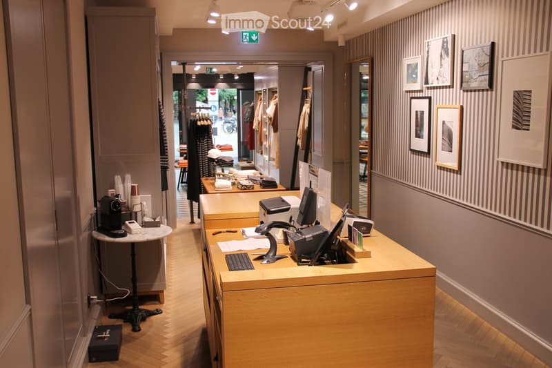 Reception area with a wooden desk, computer, coffee machine, and framed pictures on the wall. The space features a polished wooden floor and a small round table with a chair.