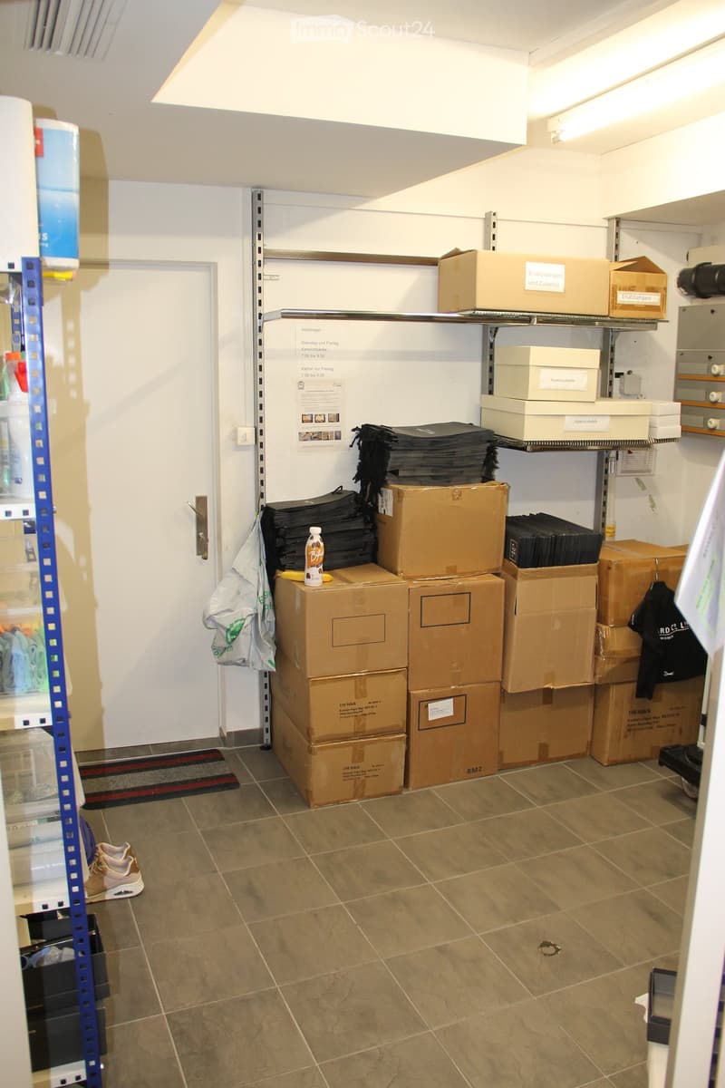 Empty storage room with tiled floor, white walls, metal shelving, and stacked cardboard boxes. A plastic bag and a pair of shoes are visible near the door.