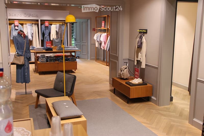 Retail store interior with wooden flooring, a small seating area with a chair and table, display racks with clothing, a mannequin, and a lamp.