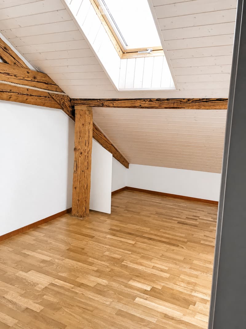 Empty attic space with wooden flooring, white walls, exposed wooden beams, and a skylight window.