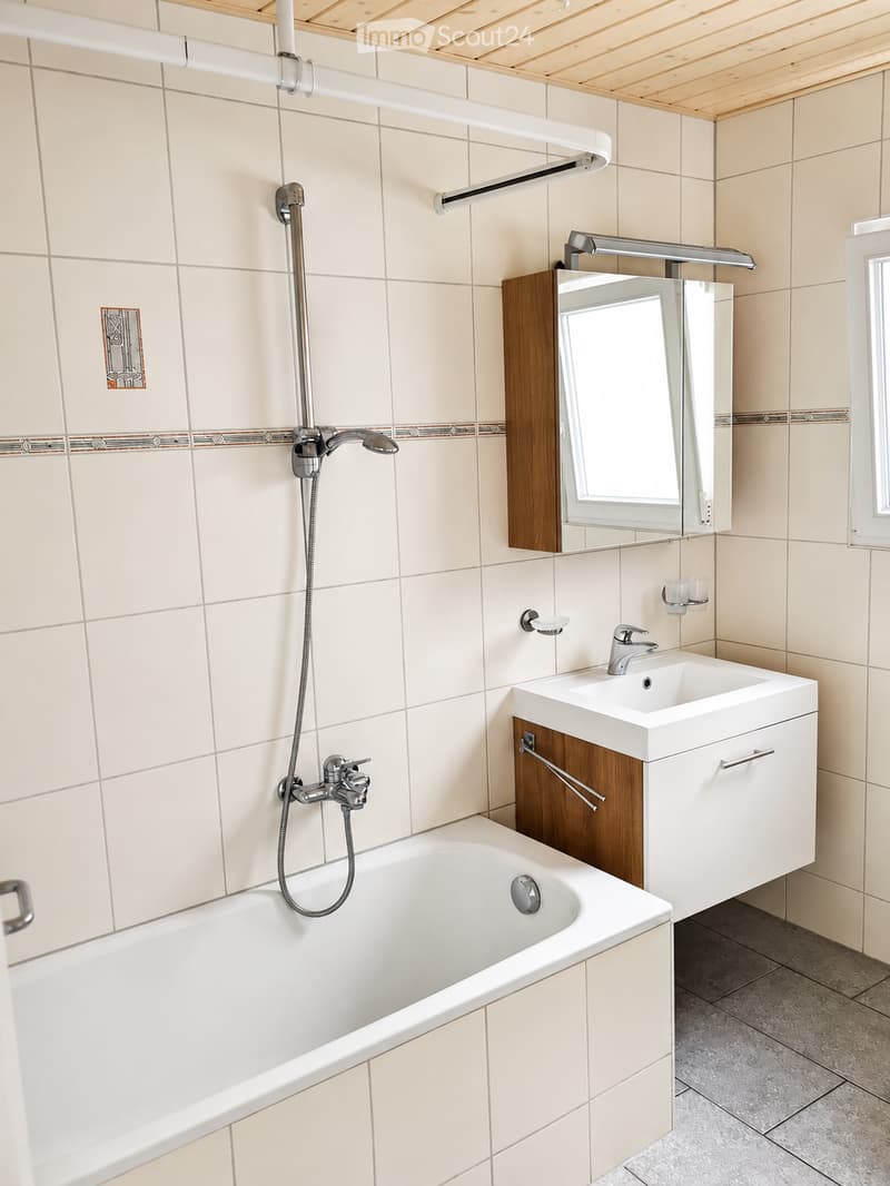 Modern bathroom with a bathtub, sink, mirror, and showerhead. Light-colored tiles and wooden accents.