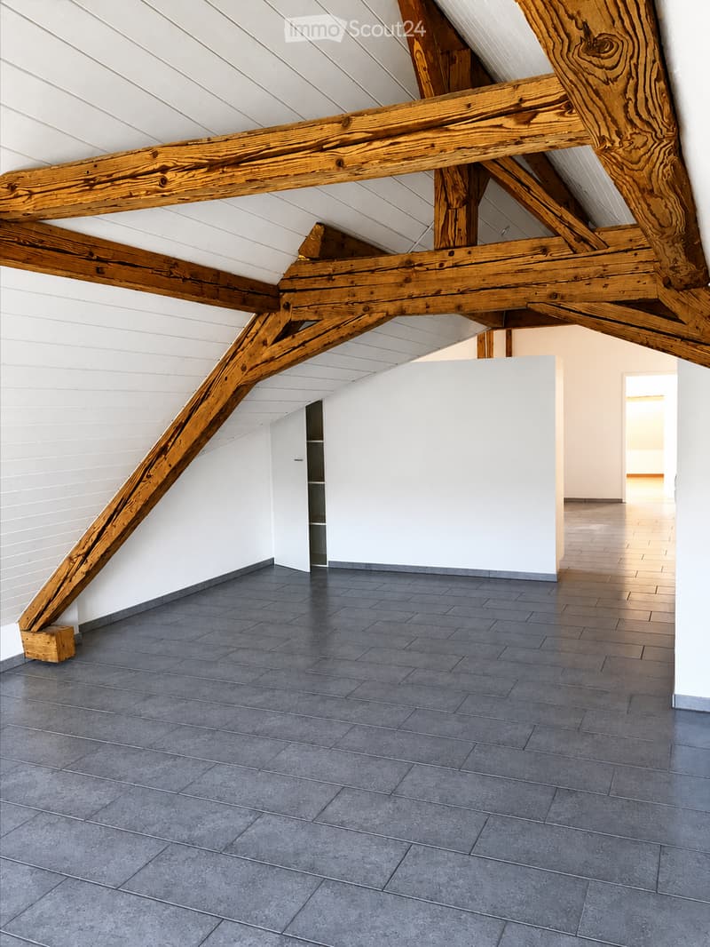 An empty attic space with exposed wooden beams, white walls, and a grey tiled floor. The room features a small built-in closet and an open doorway leading to another area.