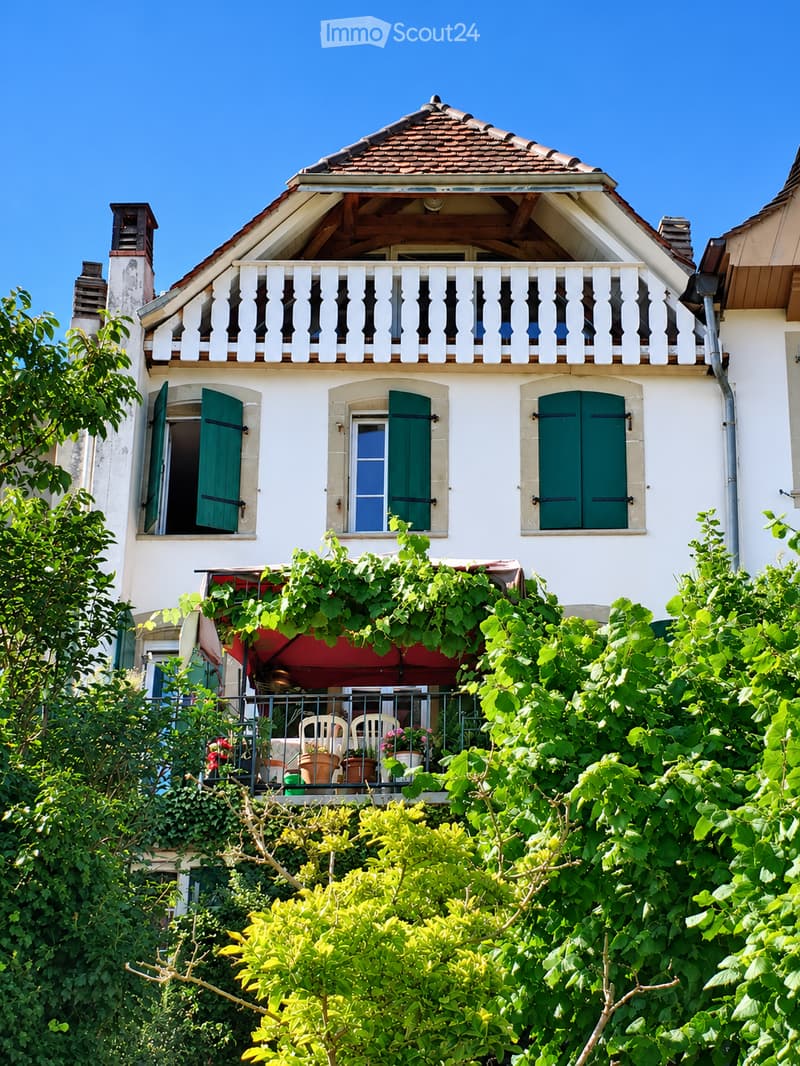 Two-story house with white exterior, green shutters, and a balcony adorned with plants and outdoor furniture.