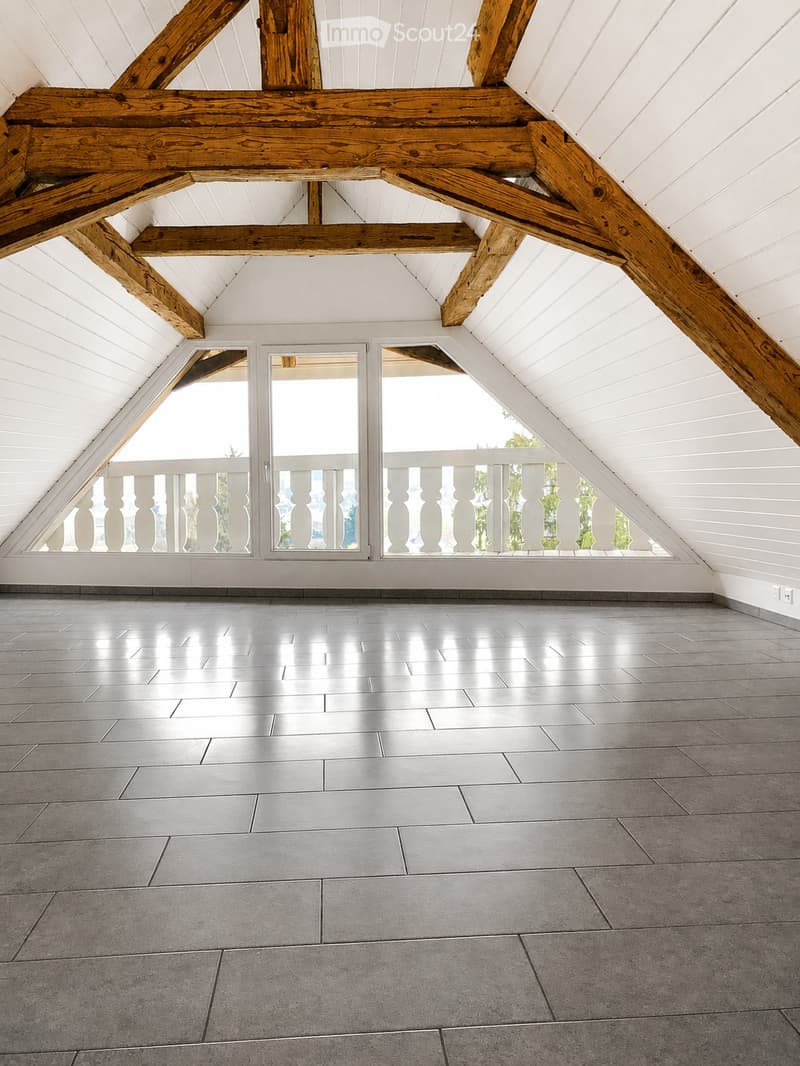 An empty attic space with exposed wooden beams, white paneled walls, and large windows with white railings, featuring a polished concrete floor.