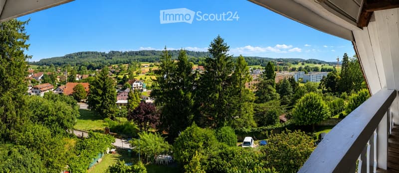 Balcony overlooking a scenic countryside with houses, trees, and distant hills under a clear blue sky.