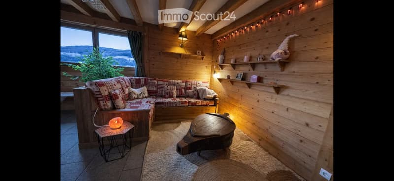 Cozy living room with wooden walls, exposed beams, large window with scenic view, plaid-patterned sofa, small coffee table, and decorative shelves.