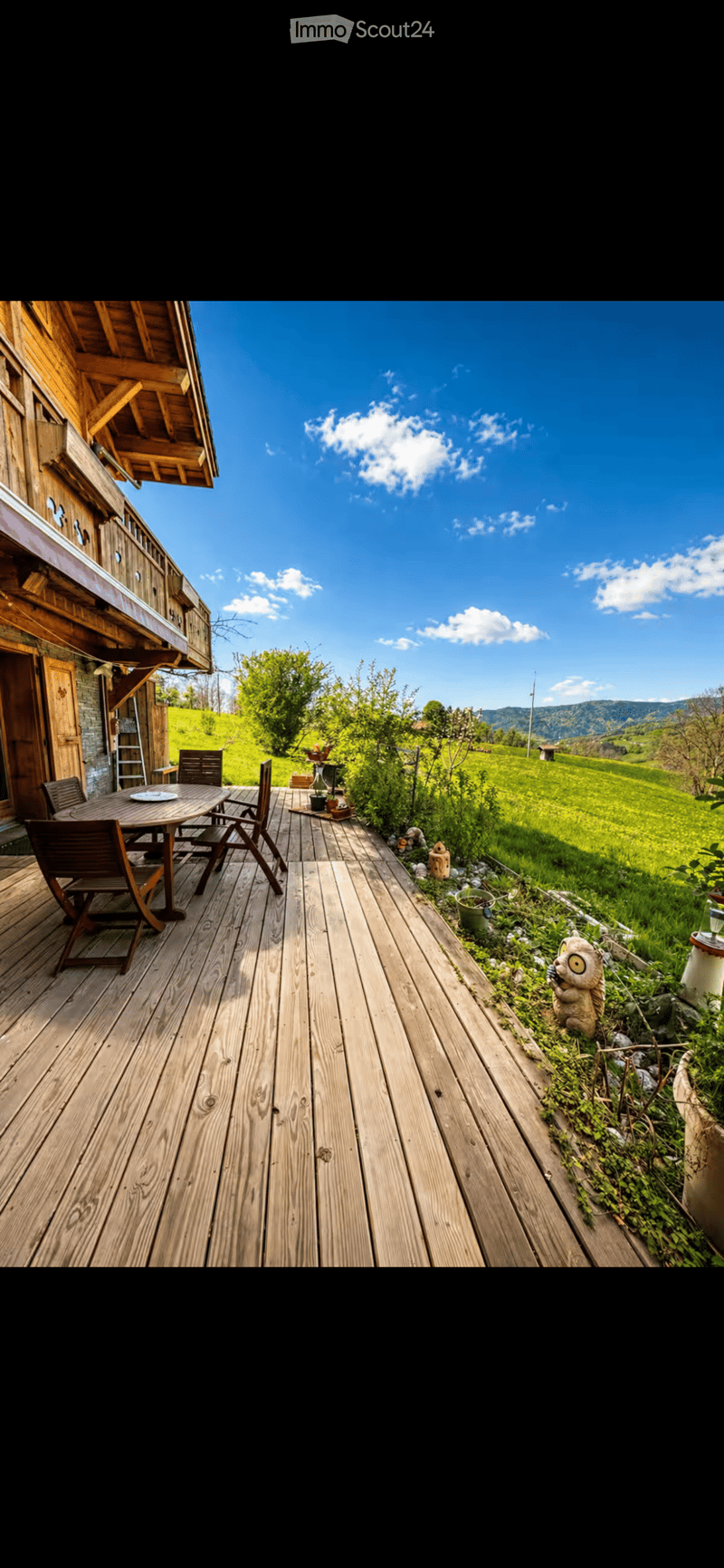 Wooden deck with a table and chairs, overlooking a green field and mountains under a clear blue sky.