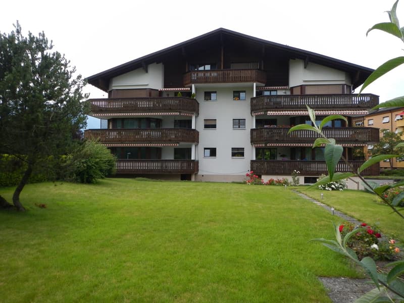 A three-story residential building with balconies, surrounded by a well-maintained lawn and garden area.