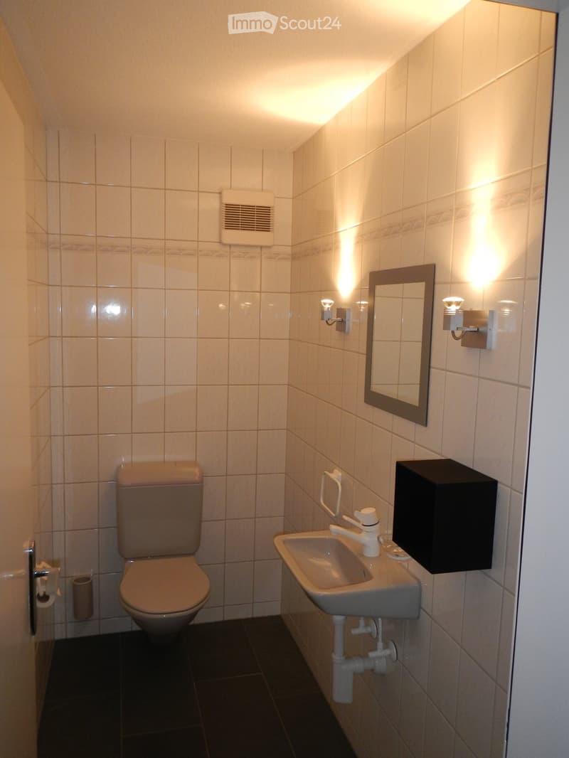Modern bathroom with beige tiles, a toilet, a sink with a mirror, and wall-mounted lighting fixtures.