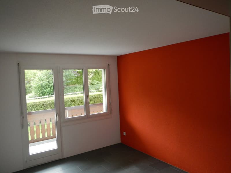 Empty room with white walls, dark tiled floor, and large windows with a view of greenery outside. The room features a white door leading to a balcony with a railing.