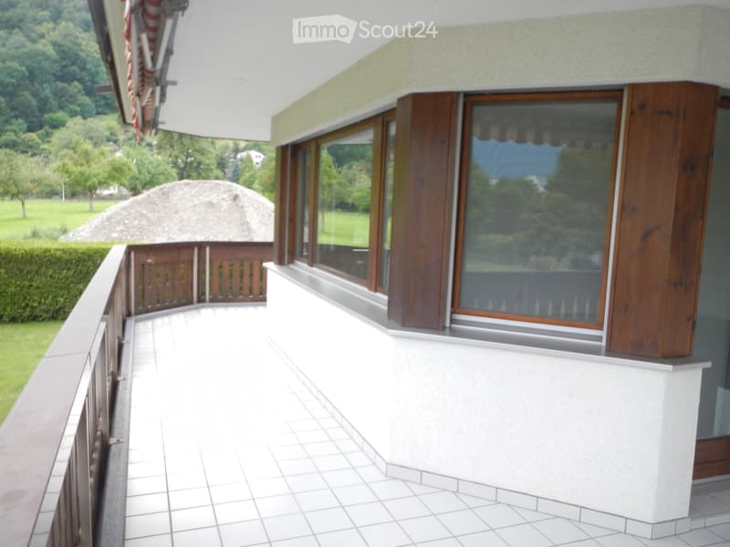 Balcony with white tiled floor, wooden railing, large windows with wooden frames, overlooking a green landscape with trees and a thatched-roof structure.
