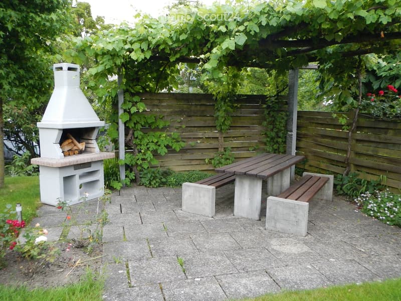 A well-maintained outdoor patio area with a stone barbecue, a wooden table with benches, and a wooden fence covered in climbing plants.
