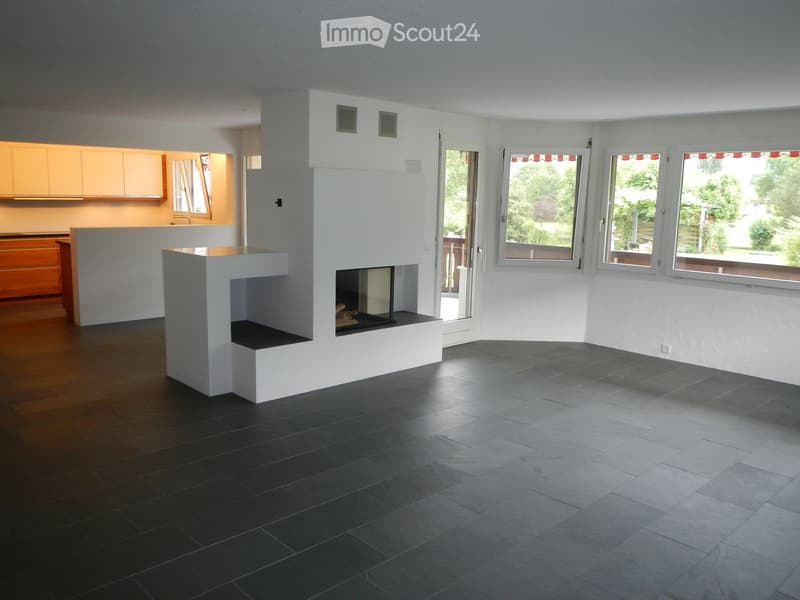 Modern living room with a central white fireplace, dark hardwood and tile flooring, large windows, and a visible kitchen area with cabinets.