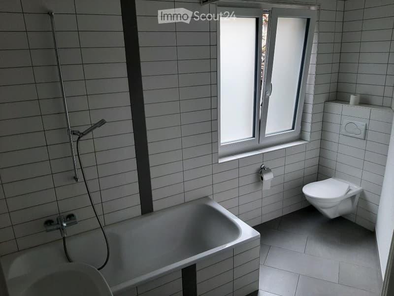 Modern bathroom with white tiled walls and floor, featuring a bathtub, toilet, and a shower hose attached to the wall. A window provides natural light.