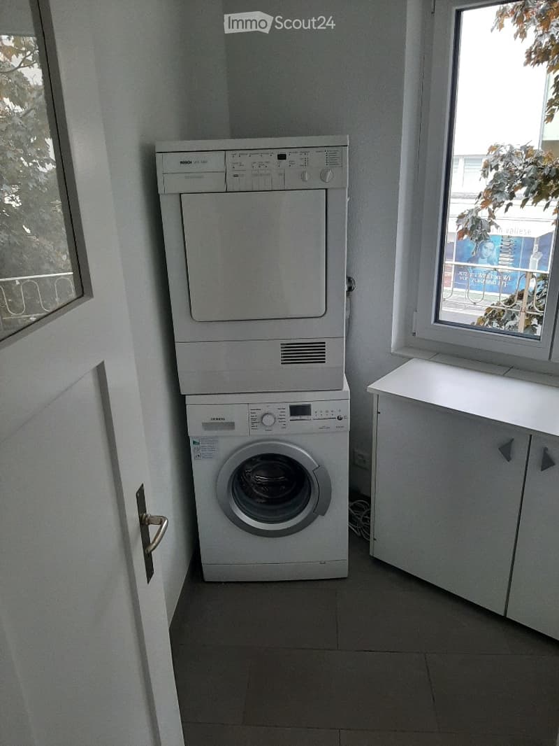 Compact bathroom with a stacked washing machine and dryer unit, white tiled walls, and a small cabinet next to the window.