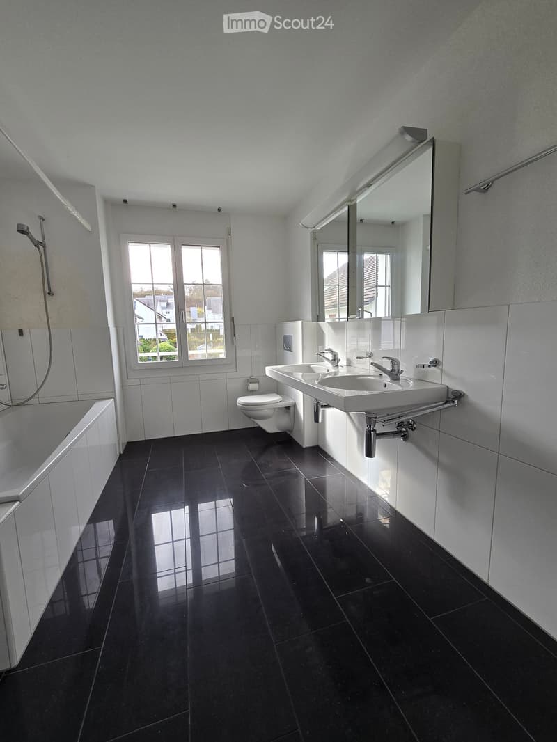 Modern bathroom with black tiled floor, white walls, bathtub, toilet, double sink, and large mirror. Natural light from a window.