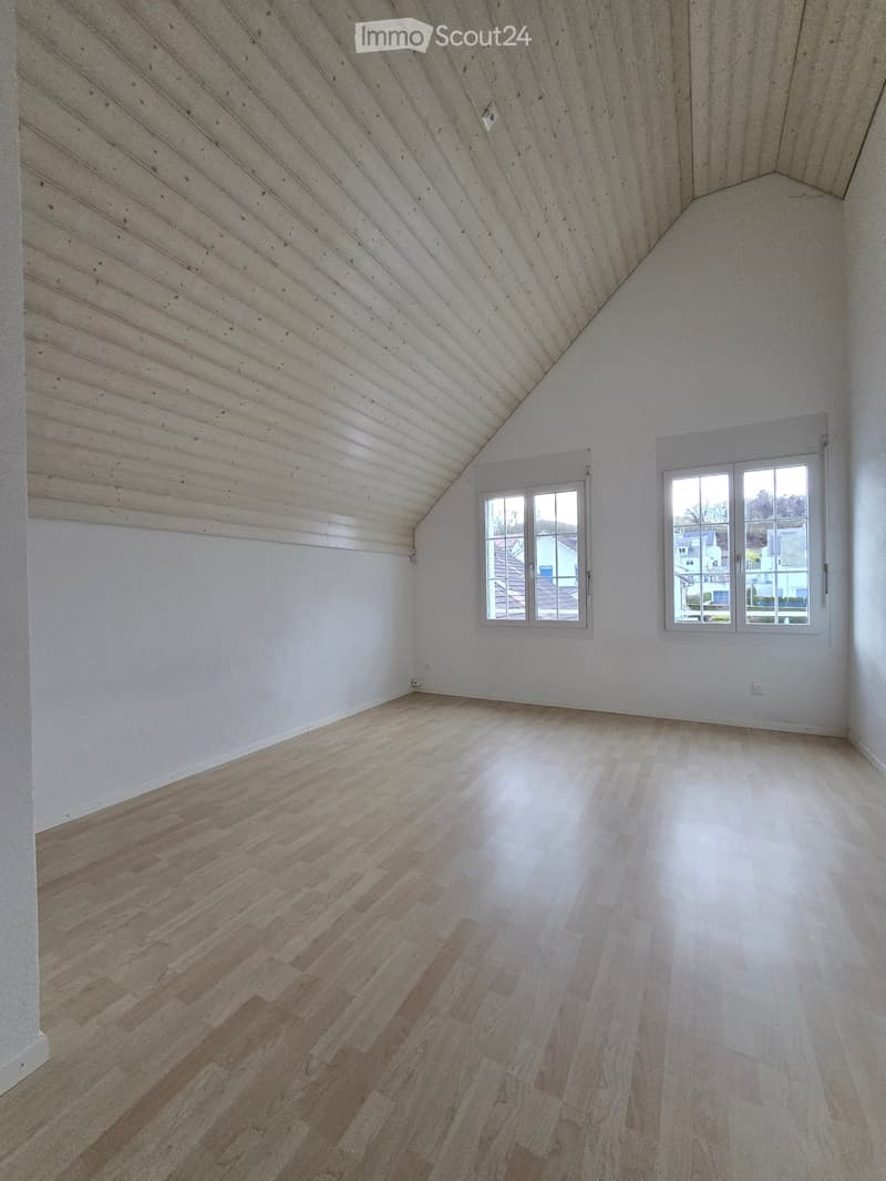 Empty room with wooden flooring and white walls, featuring two windows and a slanted wooden ceiling.