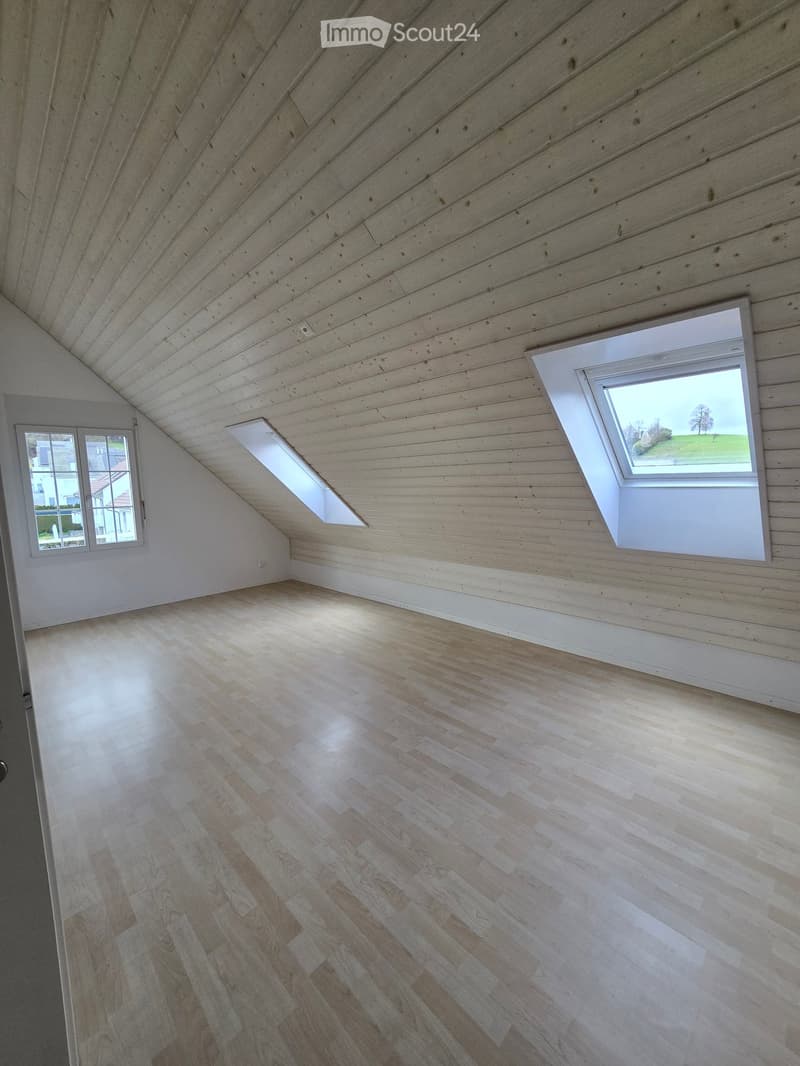 Empty attic space with wooden walls, ceiling, and floor, featuring two skylights and a window offering a view of the outdoors.