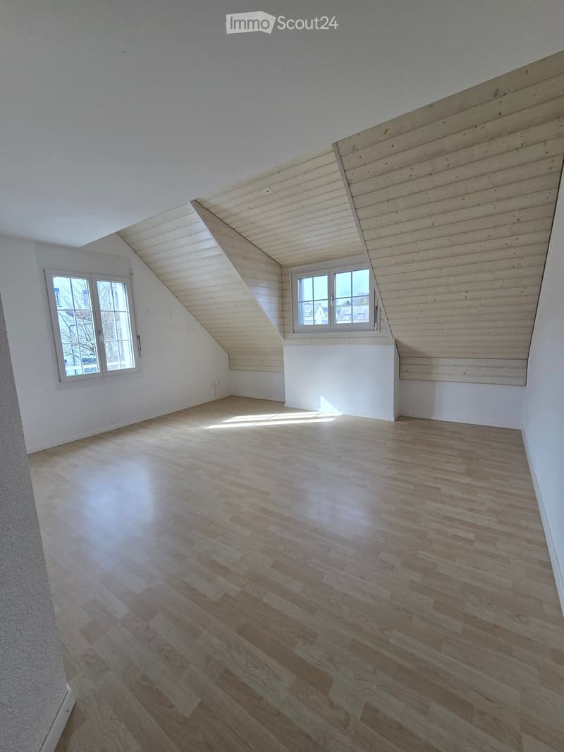 Empty attic room with wooden flooring, white walls, and two windows allowing natural light to enter.