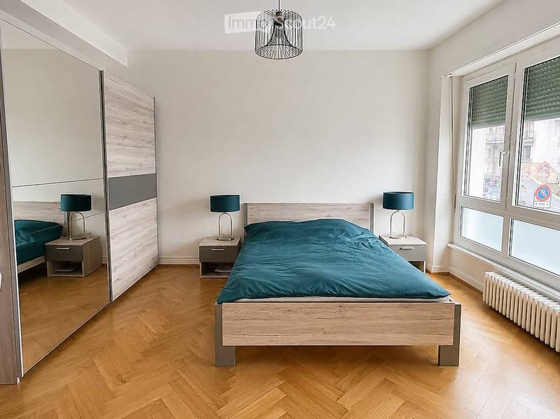 Modern bedroom with wooden flooring, a large bed with green bedding, two nightstands with lamps, and a large mirrored wardrobe.
