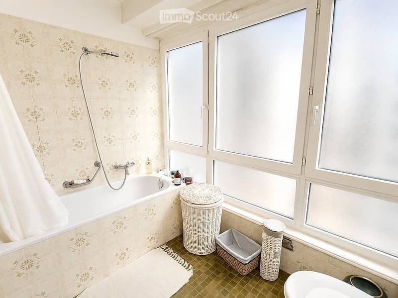 Bathroom with a bathtub, showerhead, wicker baskets, and a trash bin near large windows.