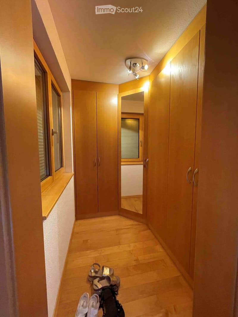 A narrow hallway with wooden flooring, built-in wardrobes, and a small window on the left side. A pair of white shoes and a pair of black shoes are placed on the floor near the camera.