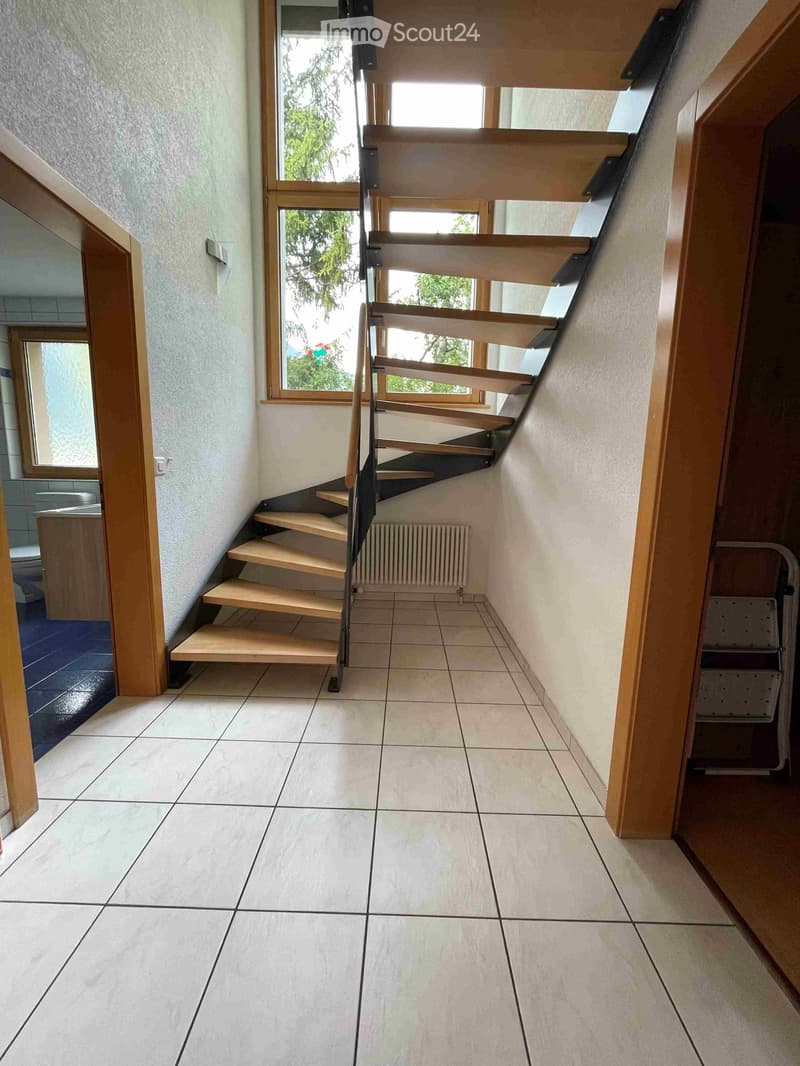 Spacious hallway with tiled floor, wooden staircase leading upwards, and natural light from windows.