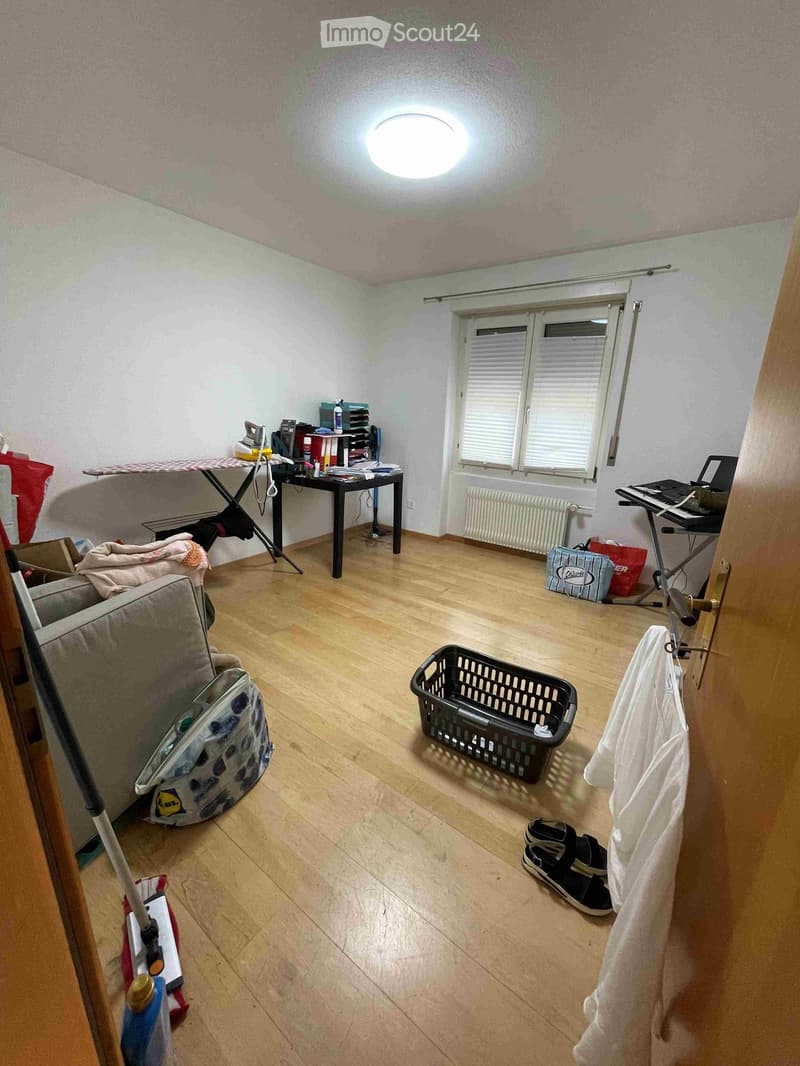 A furnished room with wooden flooring, a sofa, a table with an ironing board, a laptop, a keyboard, a basket, and a radiator with a cover.