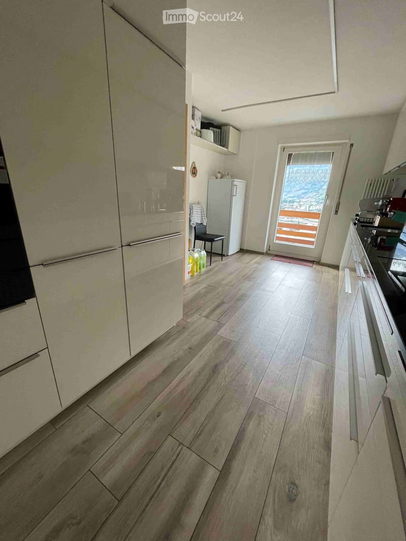 The image shows a modern, well-lit hallway with wooden flooring and white walls. There is a refrigerator, a chair, a small stool, and a built-in cupboard visible.