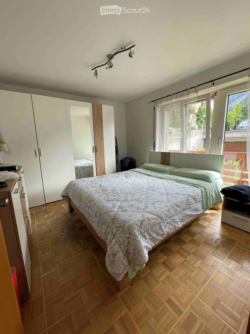 Bedroom with wooden flooring, large bed with green bedding, wardrobe with mirrored doors, nightstand with lamp, and large windows with curtains.