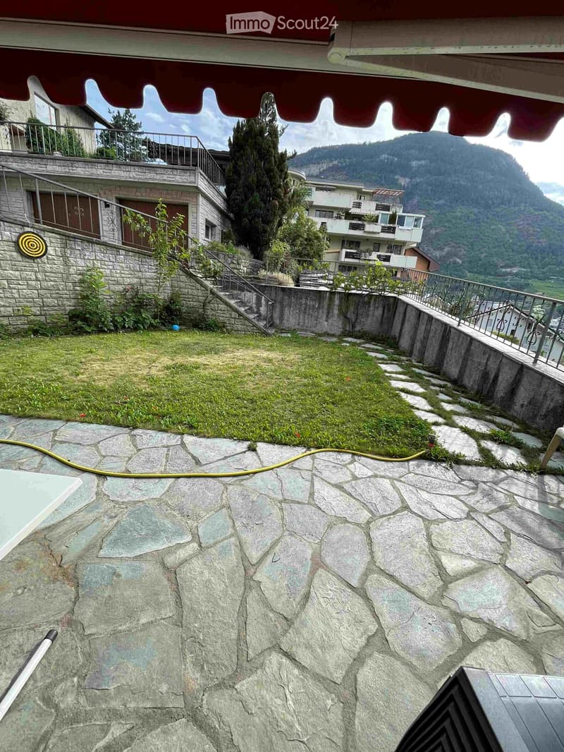 Stone-paved garden area with grassy patch, small plants, and a view of mountains and neighboring buildings.