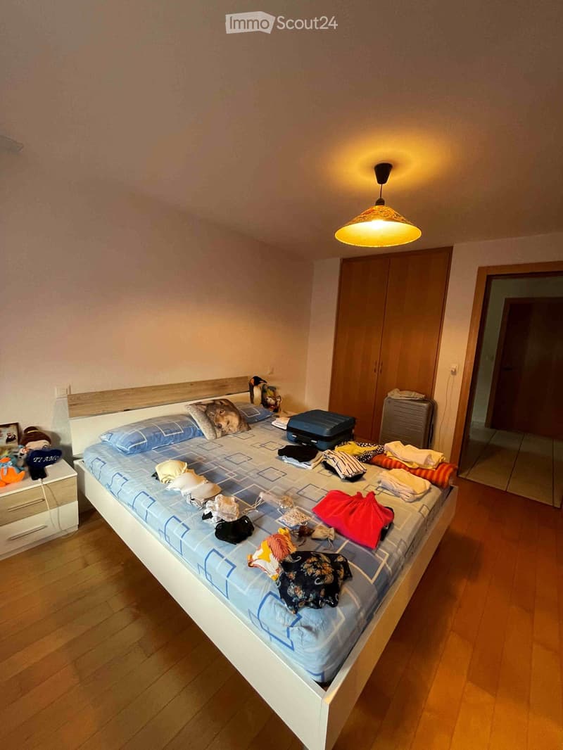 A small bedroom with a single bed, nightstand, wardrobe, and scattered clothing and items on the bed.