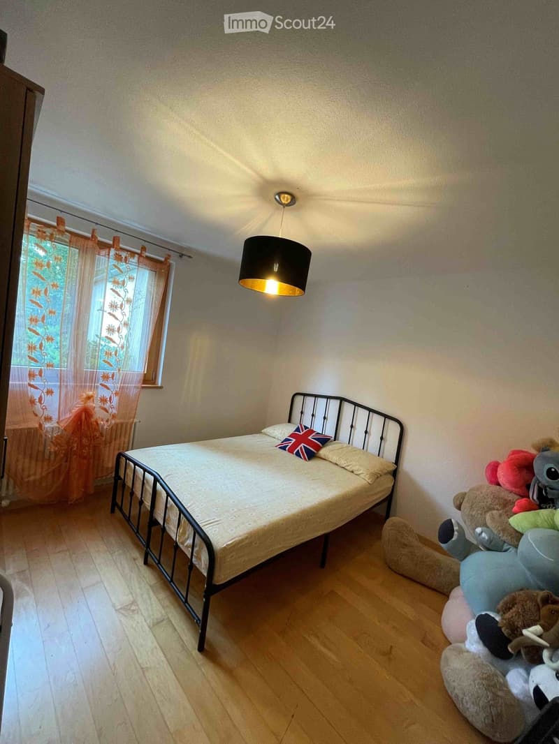 A small bedroom with a single bed, a pendant light, and a window with orange curtains. The bed has a Union Jack pillow and is surrounded by stuffed animals.