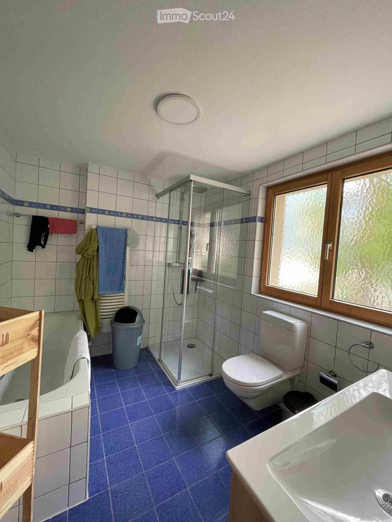 Bathroom with blue tiled floor, white toilet, sink, glass-enclosed shower, towels, and a small wooden chair.