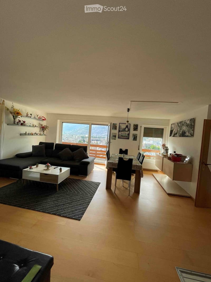 Open-plan living and dining area with wooden flooring, large windows offering a scenic view, a black sectional sofa, a white coffee table, a dining table with chairs, and decorative shelves.