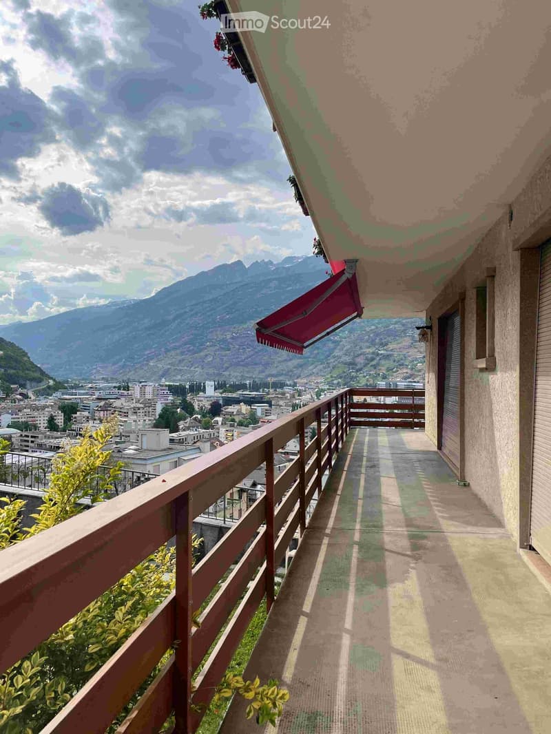 Balcony with wooden railing, overlooking a cityscape and mountains, with a red awning and some greenery visible.