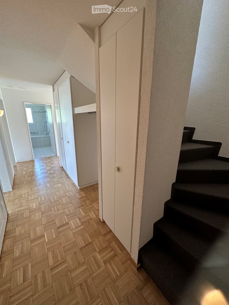 A modern hallway with wooden herringbone flooring, white walls, and a staircase leading upwards. A closet with mirrored doors is visible on the left, and a bathroom is partially visible at the end of the hallway.