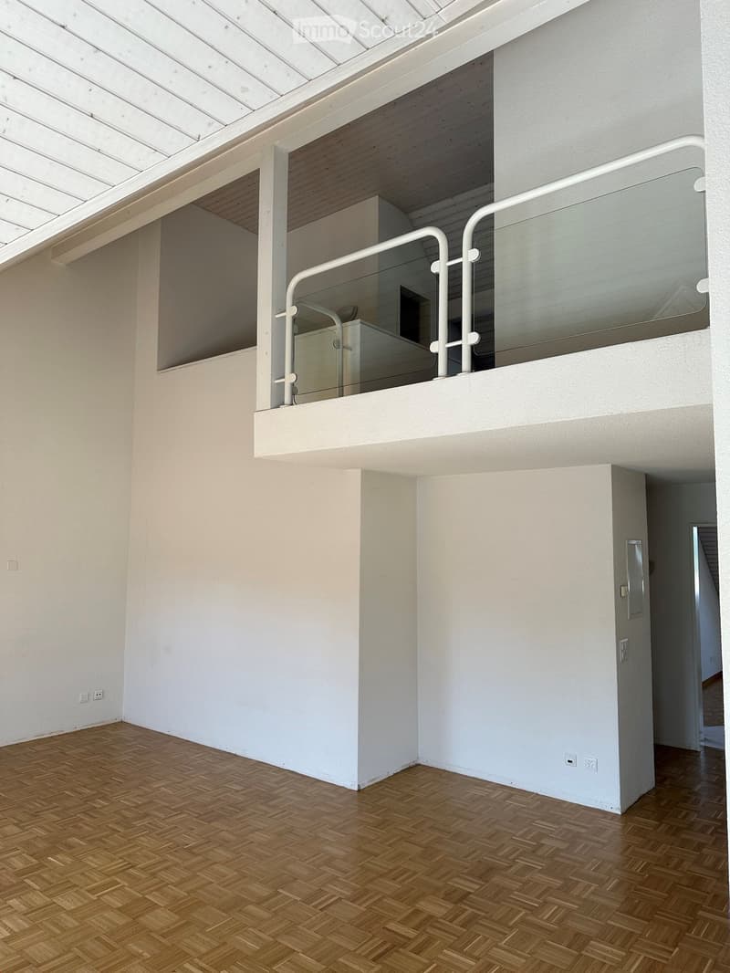 An empty room with wooden herringbone flooring, white walls, and a glass-enclosed balcony area with white railings. The ceiling has a skylight, and there are electrical outlets visible on the walls.