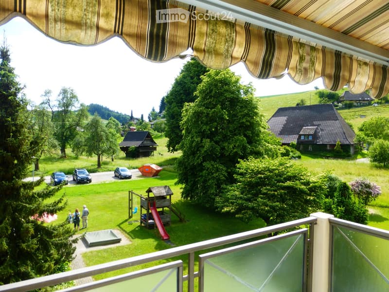 View from a balcony showing a well-maintained garden with a playground, slide, swing, and several parked cars. The area is surrounded by trees and hills with houses in the background.