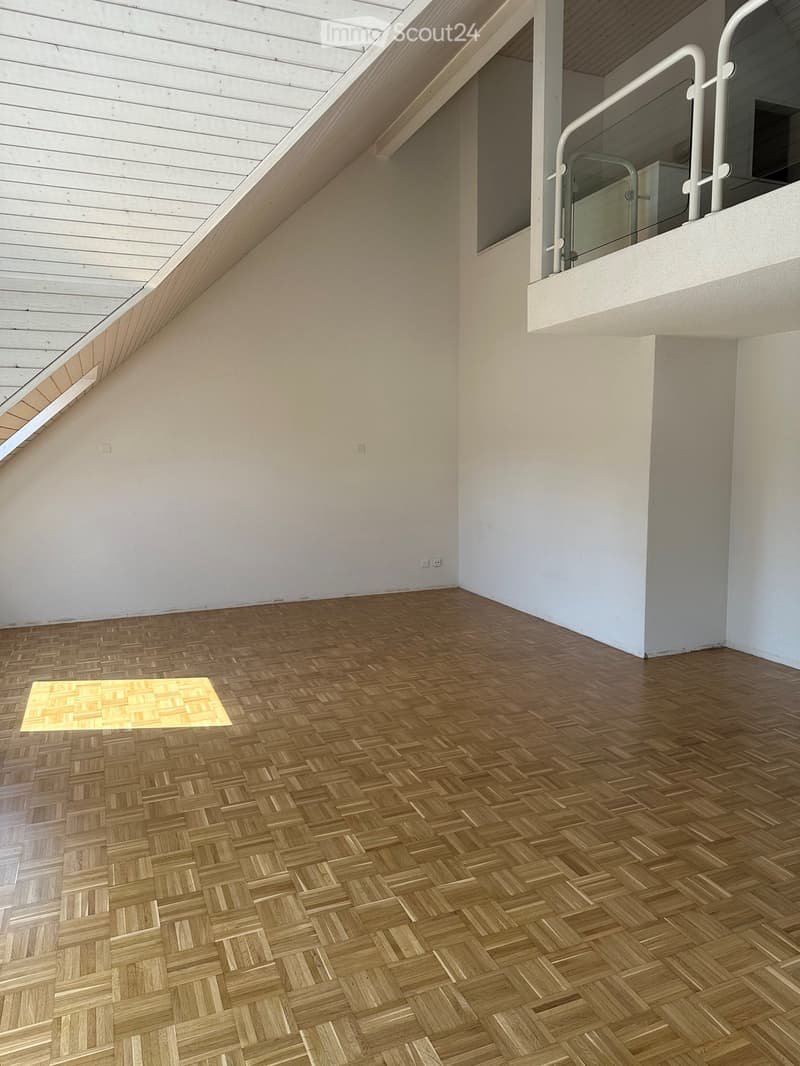Empty room with wooden herringbone flooring, white walls, and a small balcony with glass railing visible in the background.