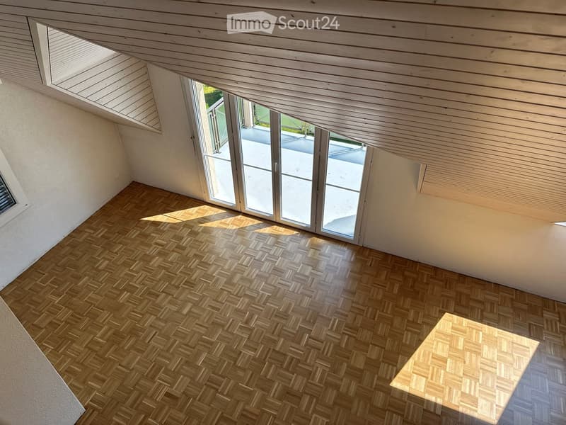An unfurnished room with wooden herringbone flooring, white walls, and large sliding glass doors leading to an outdoor area. The ceiling is slanted with skylights.