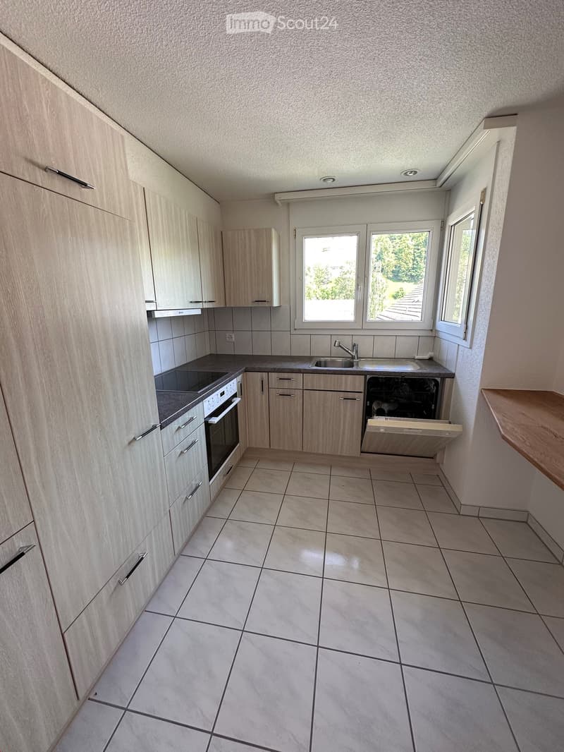 Modern kitchen with light wood cabinets, tiled floor, oven, sink, and dishwasher. Large windows provide natural light.