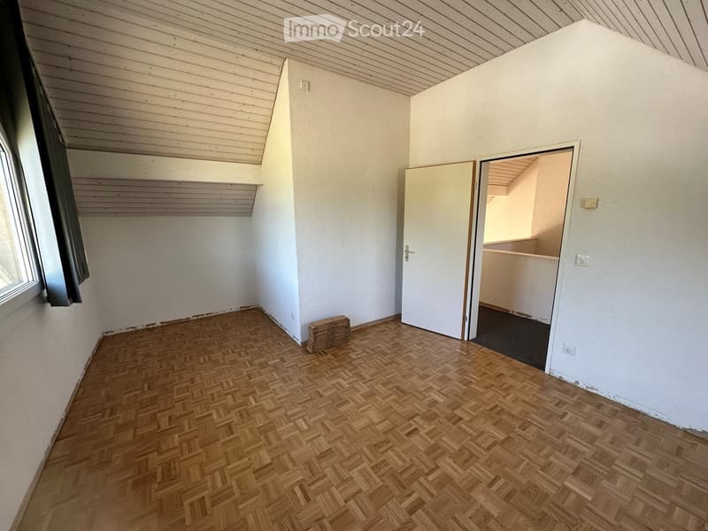 Empty room with wooden herringbone flooring, white walls, and a slanted wooden ceiling. A door leads to another room, and a small stack of wooden planks is visible near the wall.