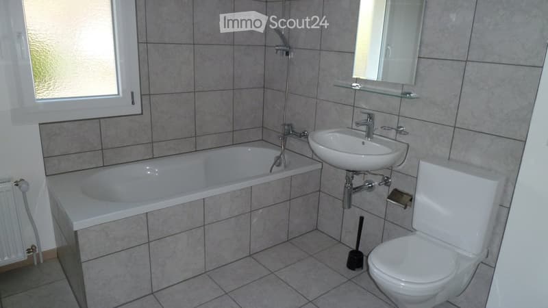 Modern bathroom with a bathtub, sink, toilet, mirror, and radiator. The walls are tiled, and there is a window providing natural light.