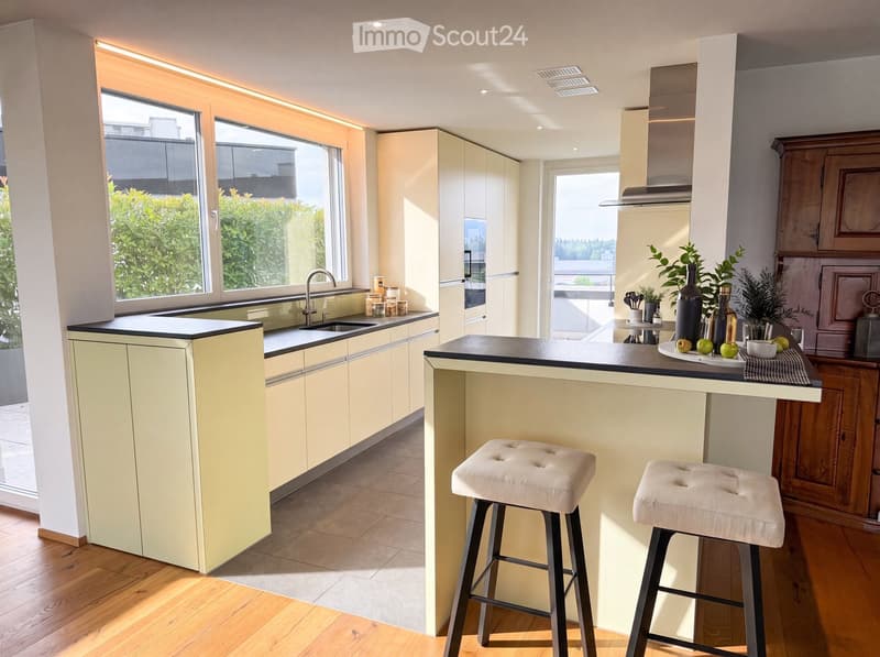 Modern kitchen with light-colored cabinets, a central island with bar stools, and a sink area. The space features a sleek design with wooden flooring and ample natural light from large windows.