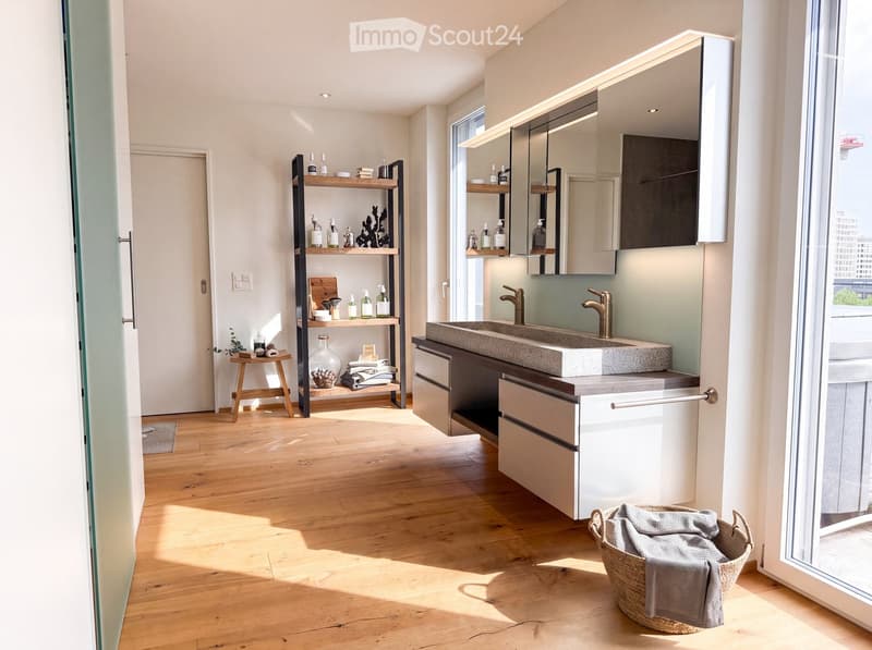 Modern bathroom with wooden flooring, a large sink with a modern faucet, a mirrored cabinet, a wooden shelf with toiletries, a small wooden stool, and a woven basket with a towel.