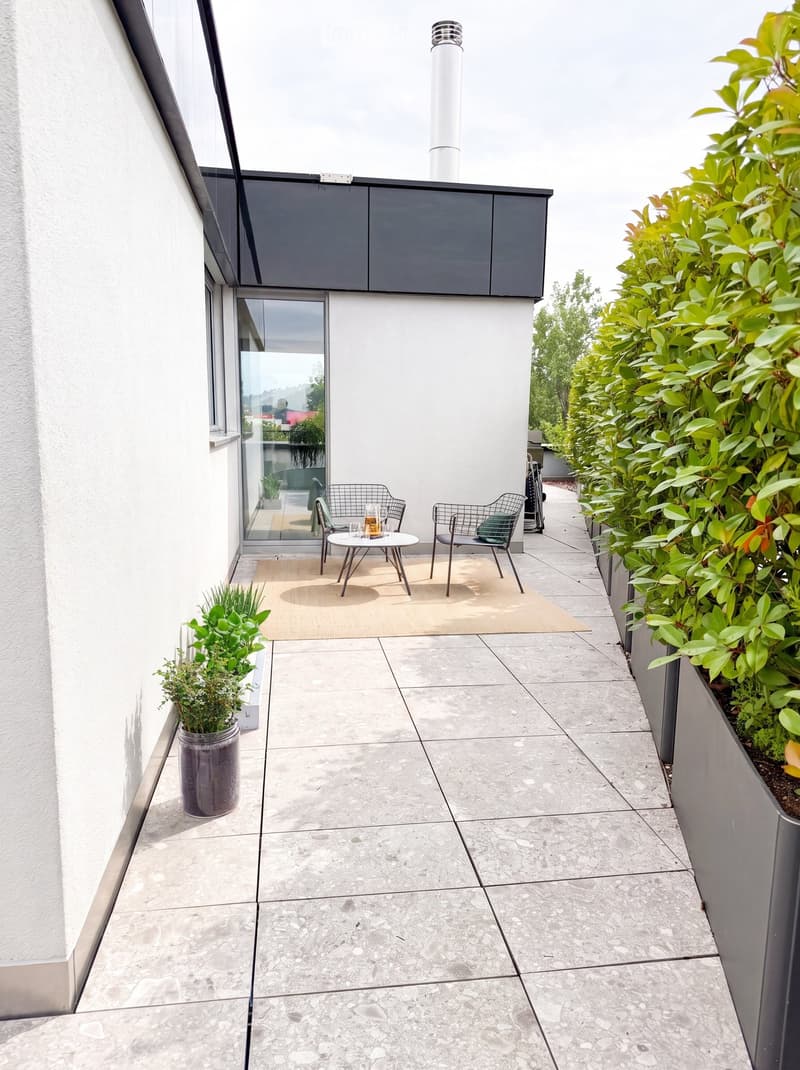 Modern balcony with grey tiled floor, white walls, small seating area with a table and two chairs, surrounded by potted plants and greenery.