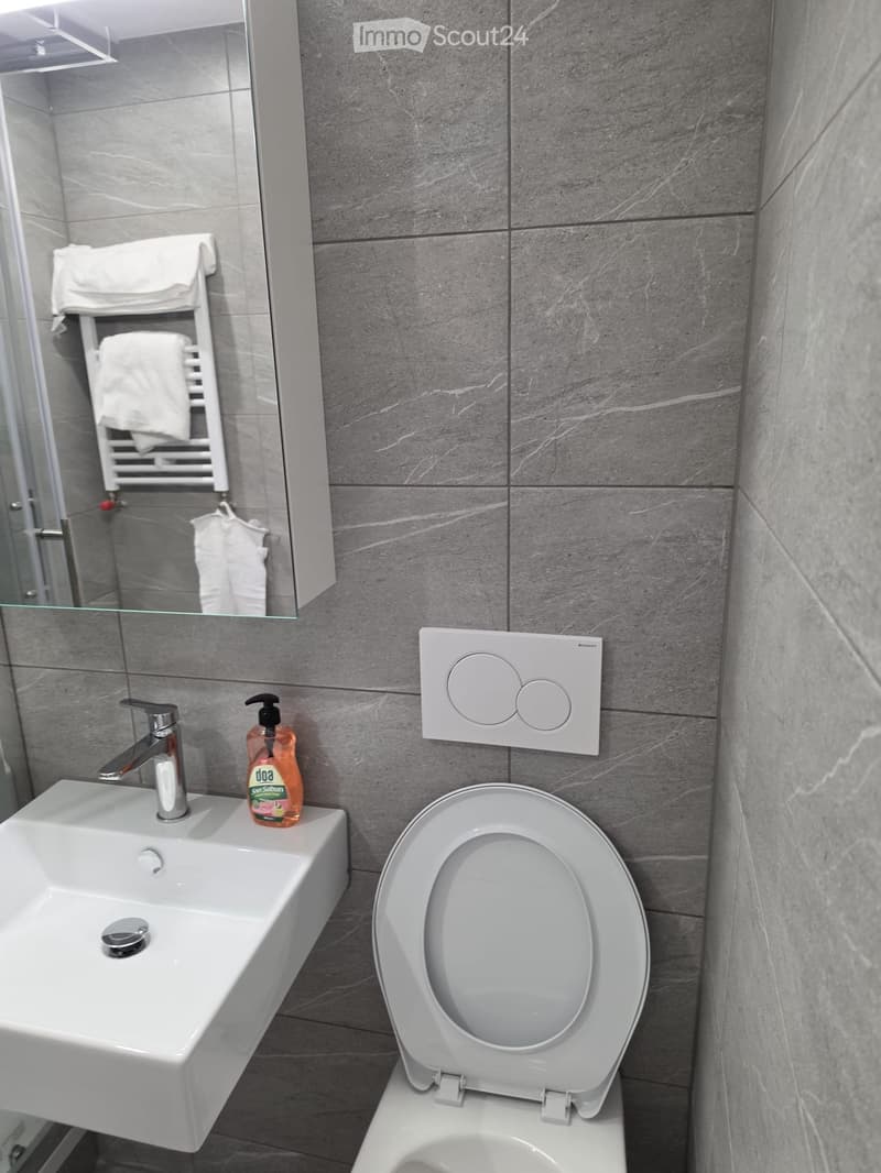 Modern bathroom with a white sink, chrome faucet, soap dispenser, and a toilet with the lid up. Grey marble tiles on the walls and a towel rack with white towels in the background.