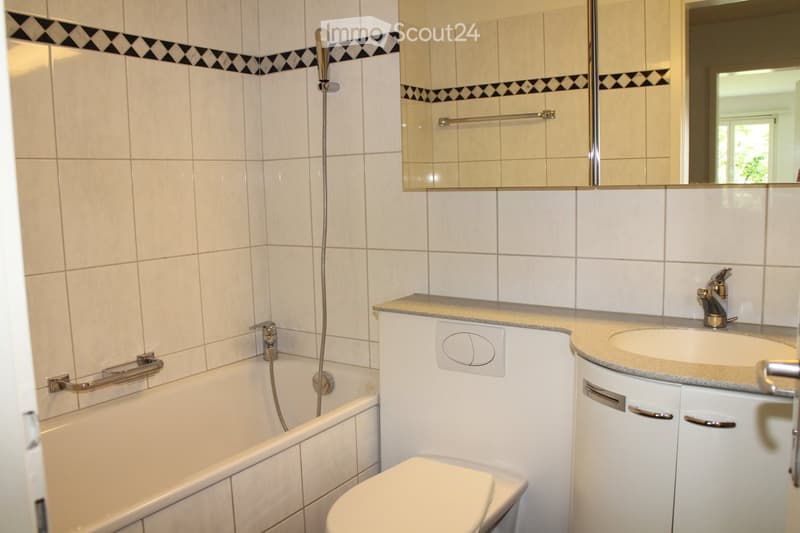 Bathroom with a bathtub, toilet, sink, mirror, and showerhead. Features tiled walls with a decorative black and white border, a countertop with a sink, and a handrail for safety.