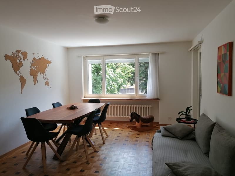 Bright living room with wooden flooring, large windows, a sofa, a dining table with chairs, a small plant, and a decorative world map on the wall.